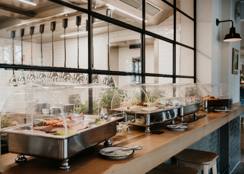 Frühstücksbuffet mit Chafing Dishes unter Glashauben auf einer Theke aus Holz und schwarzem Stahl