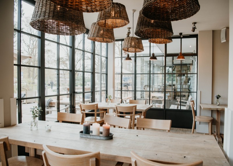Restaurant mit langer Holztafel, Korbleuchten und schwarzen Sprossenfenstern zum Park