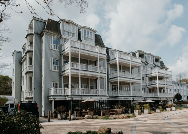 Hotelgebäude mit hellgrauer Holzfassade und umlaufenden weißen Balkonen, Terrasse davor