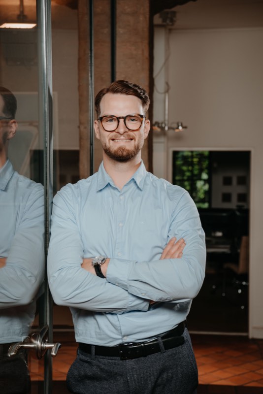 Businessporträt: Mann mit Brille im hellblauen Hemd, Arme verschränkt, an einer Glastür
