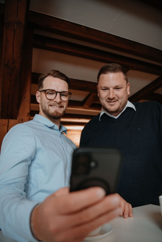 Zwei Kollegen schauen gemeinsam auf ein Smartphone, alte Dachbalken darüber