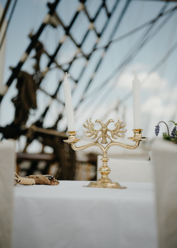 Goldener Kerzenleuchter auf weißer Tafel an Deck, dahinter die Takelage