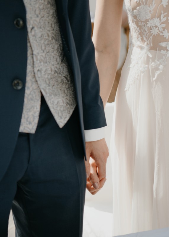 Braut und Bräutigam halten Händchen, Spitzenkleid neben dunklem Anzug mit Weste