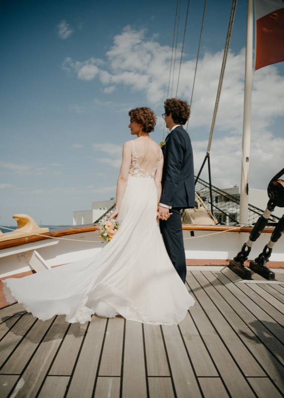 Braut im fließenden Kleid mit Brautstrauß auf dem Holzdeck, der Bräutigam an ihrer Seite