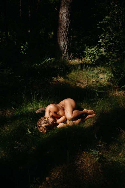 Aktaufnahme im Wald: eine Figur liegt eingerollt in einem Sonnenfleck auf dem Moos