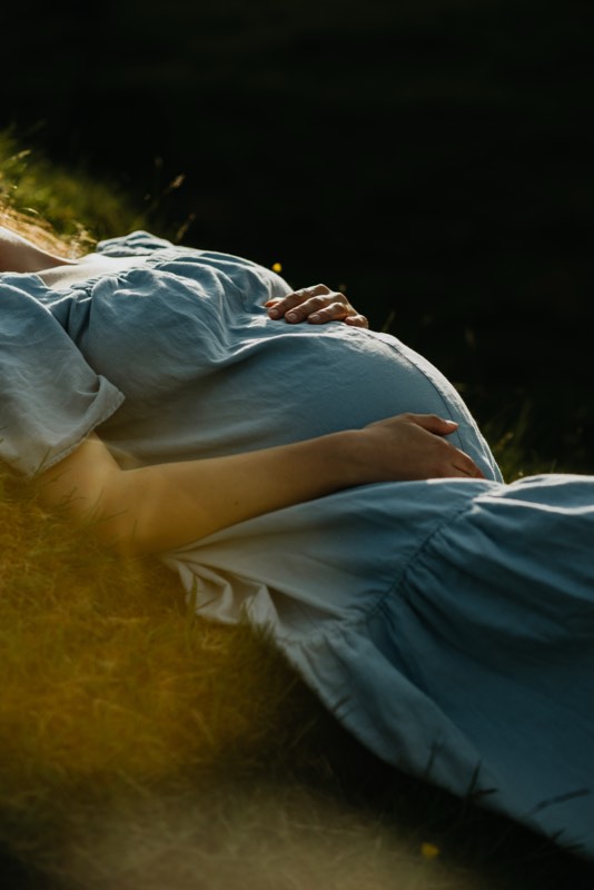 Nahaufnahme des Babybauchs im hellblauen Kleid, beide Hände darauf, Abendlicht